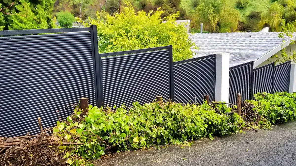 Dark aluminum slat fencing stepping down a hillside above a road, with planting along its base