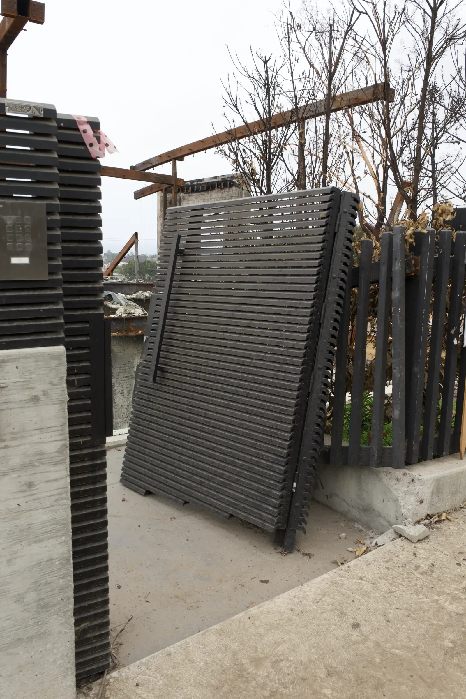 A Mulholland aluminum slat gate panel standing upright amid the burned frame of a destroyed house