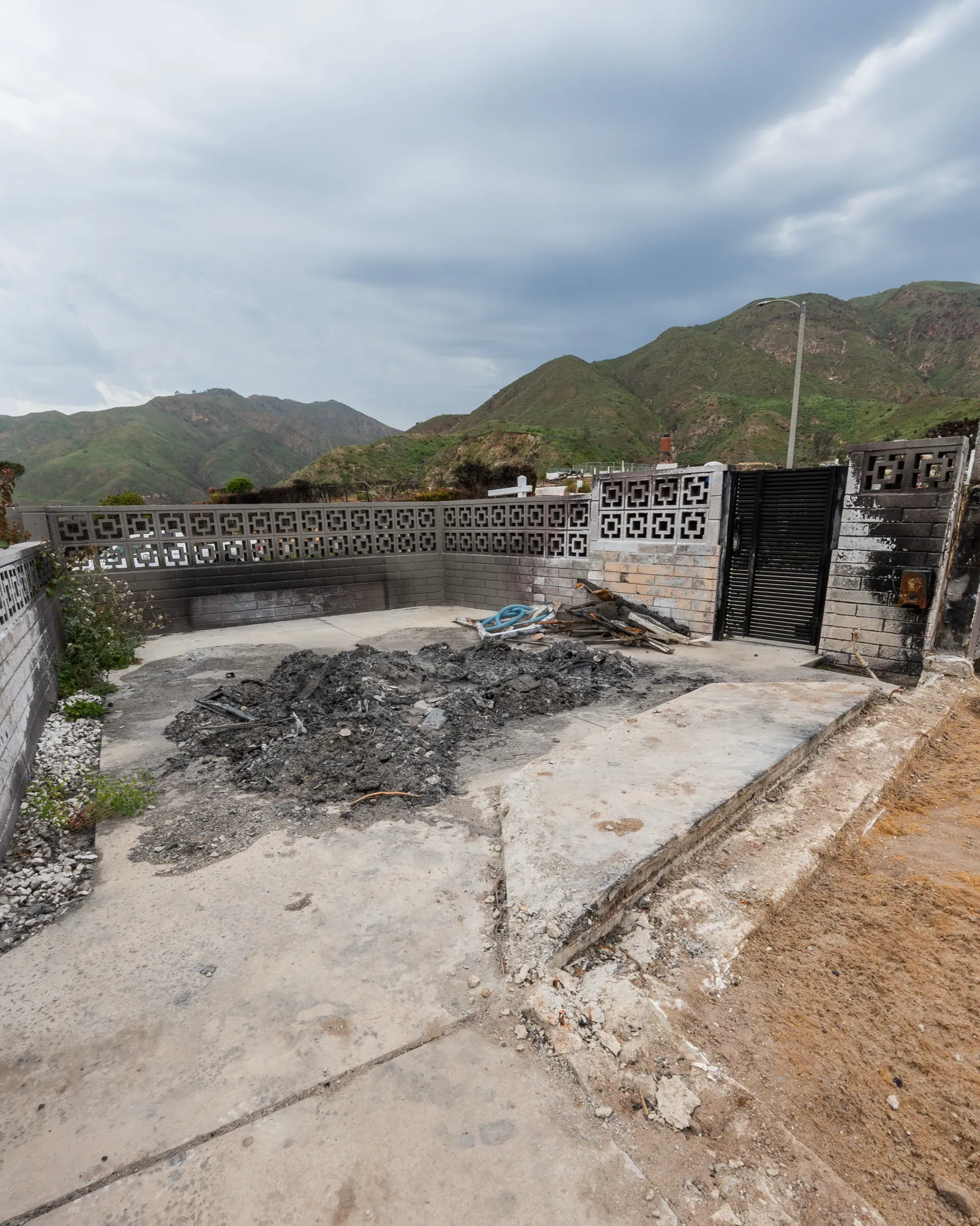 A black Mulholland gate and perimeter wall standing intact on a property burned down to its concrete slab, hills behind