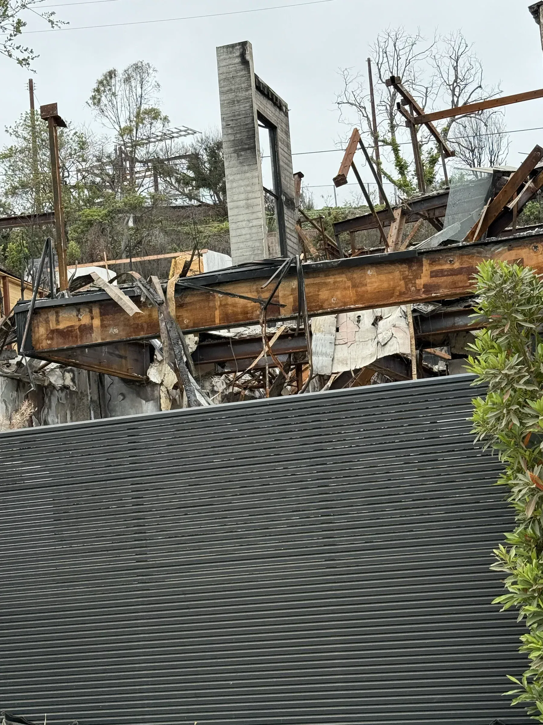 A grey Mulholland slat fence standing clean in front of a collapsed, fire-damaged structure