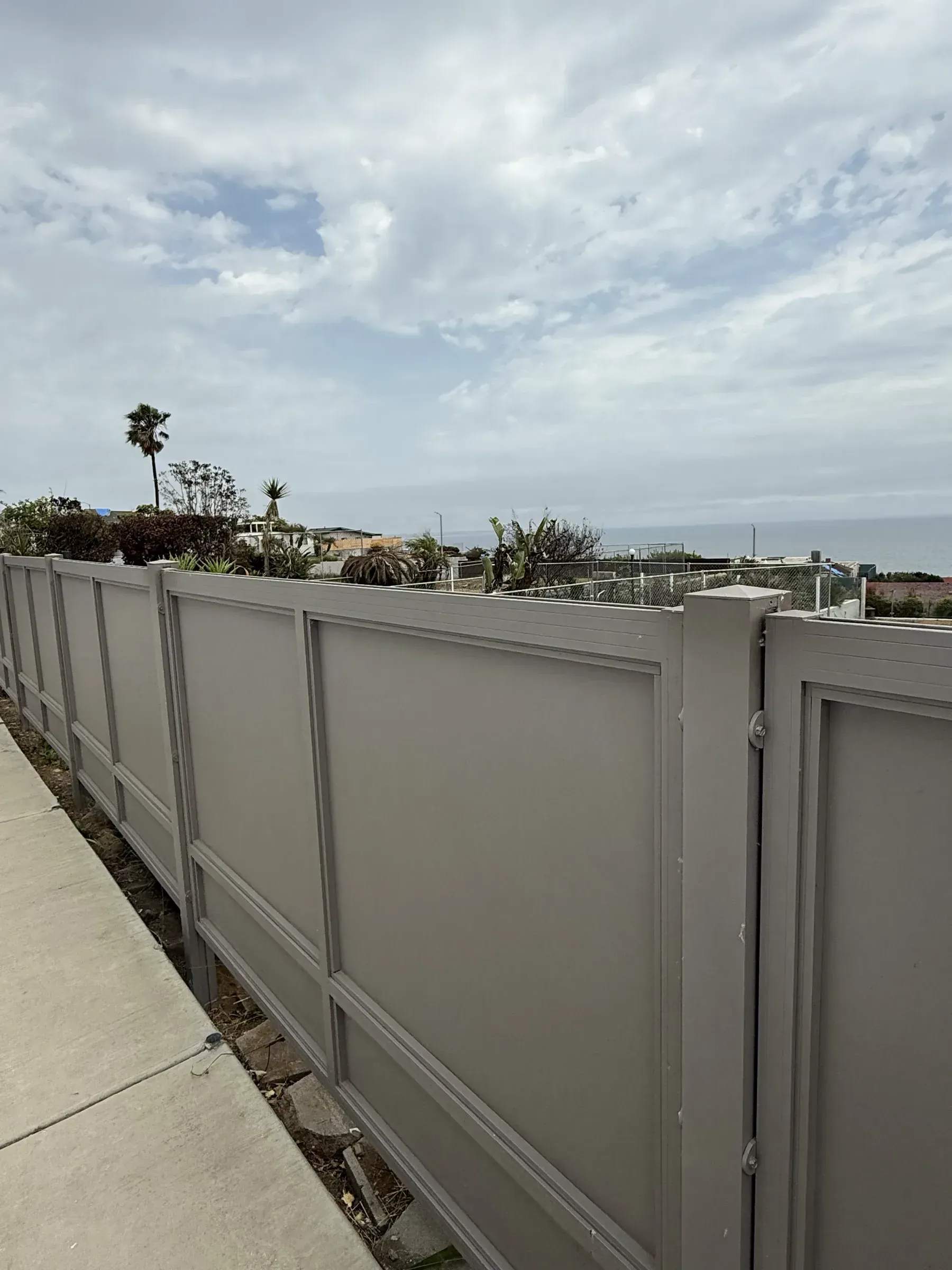 A grey Mulholland solid-panel aluminum fence running along a coastal bluff above the ocean