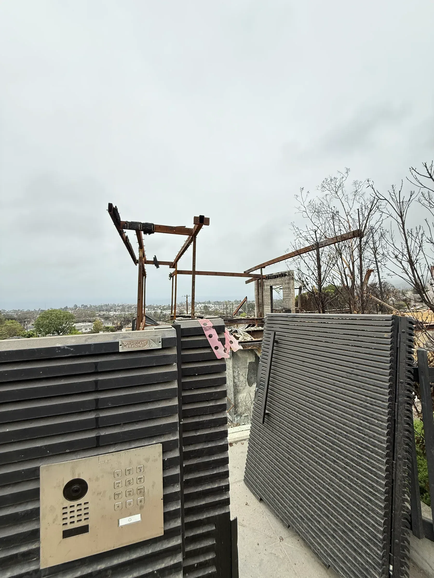 An undamaged Mulholland gate and video intercom in front of the burned steel frame of a hillside house, ocean beyond