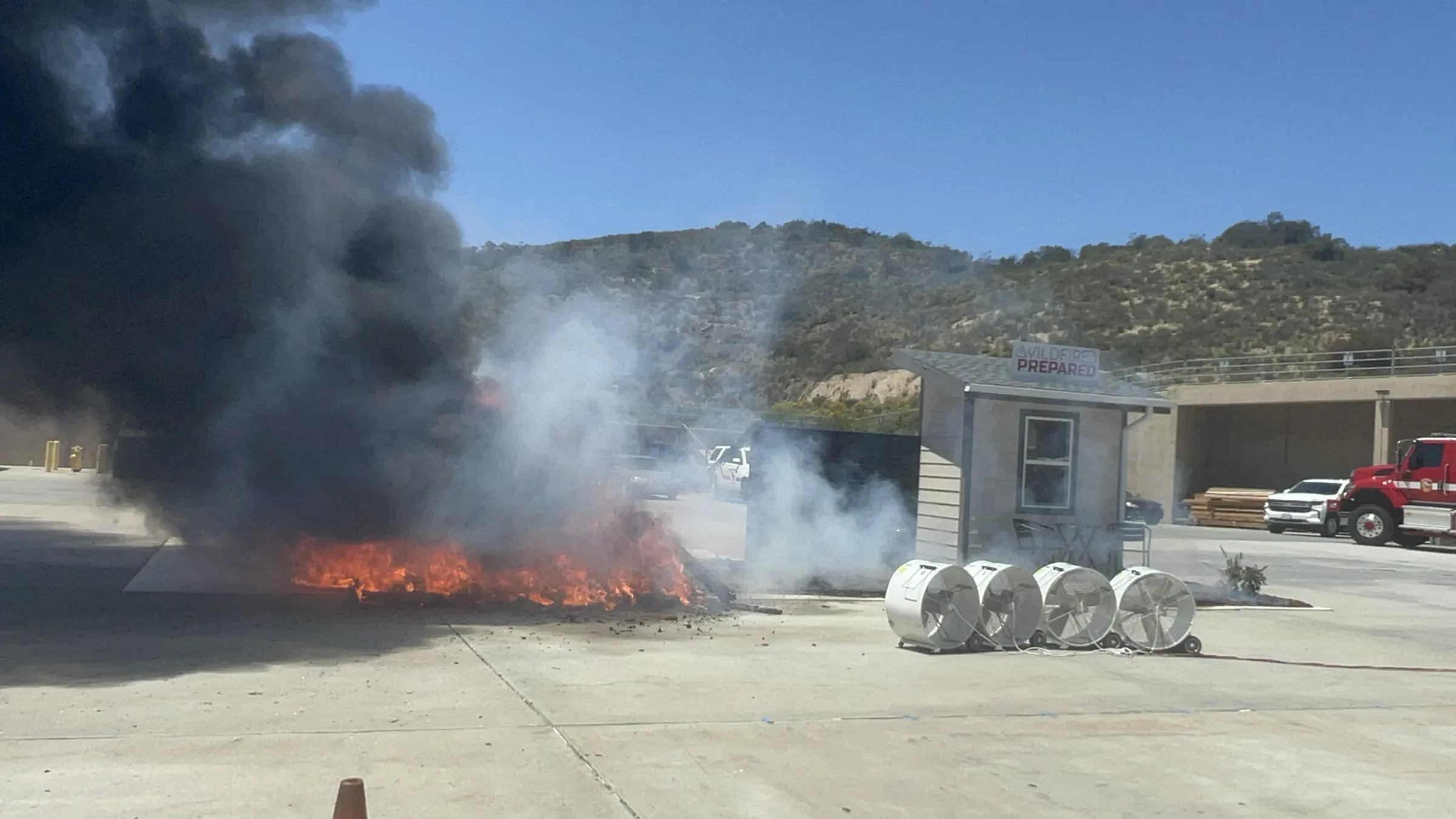 Wind-driven fire burning at full height next to the test structure, wind machines and fire crew standing by