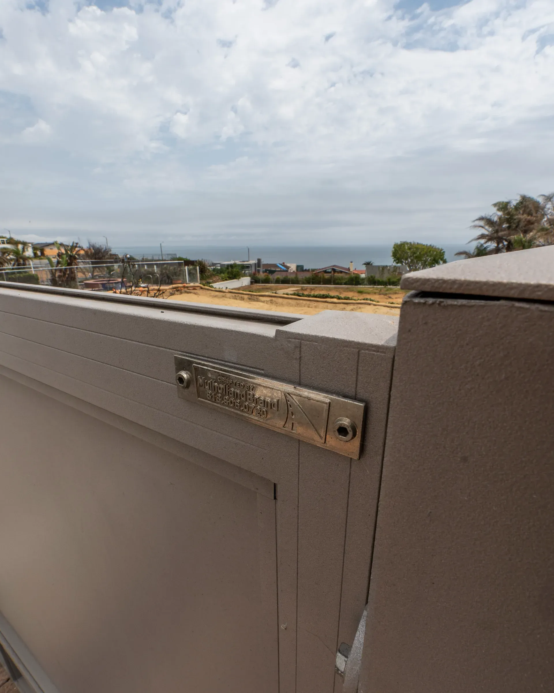 The Mulholland maker's plate on a grey aluminum gate overlooking cleared coastal lots and the ocean