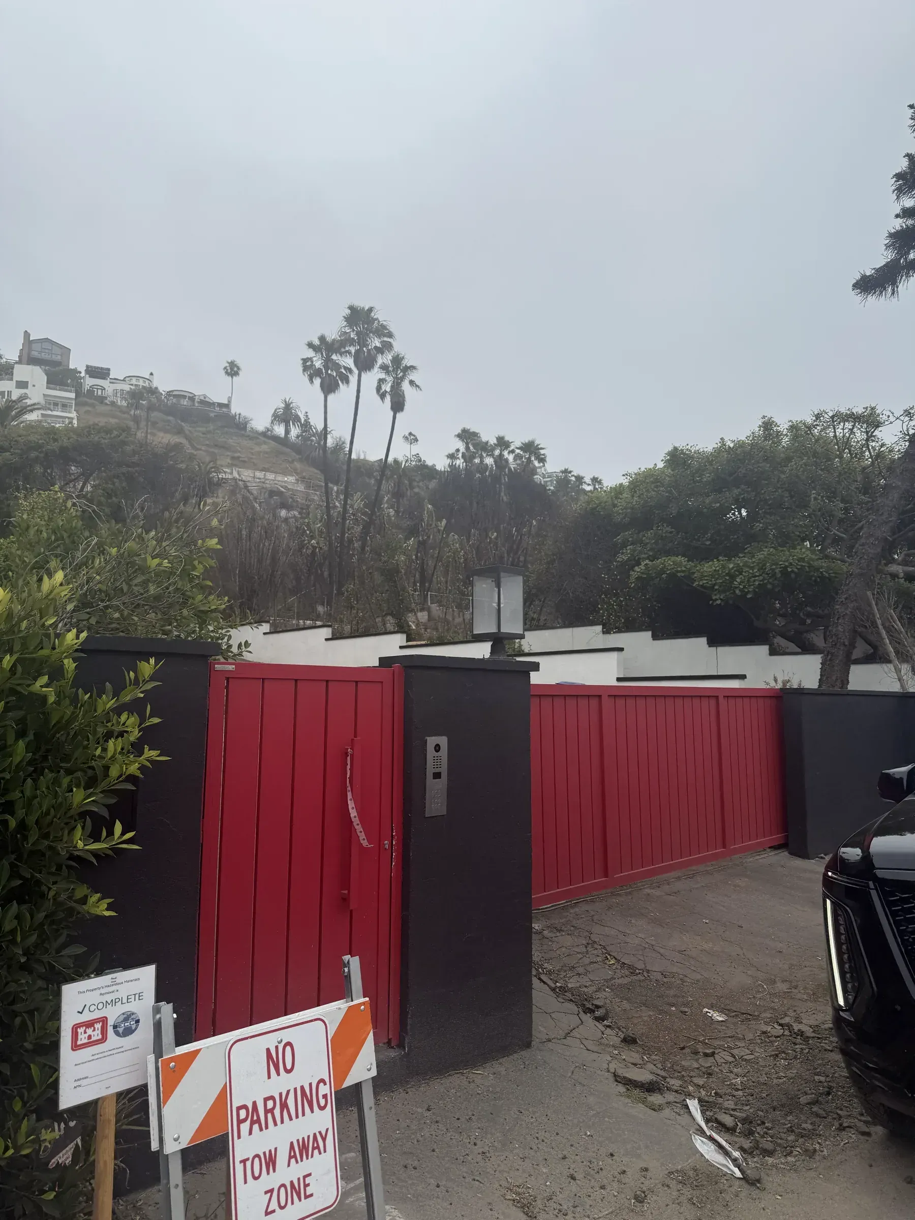 A red Mulholland driveway gate and matching fence at a hillside property with palms and rebuilt homes above