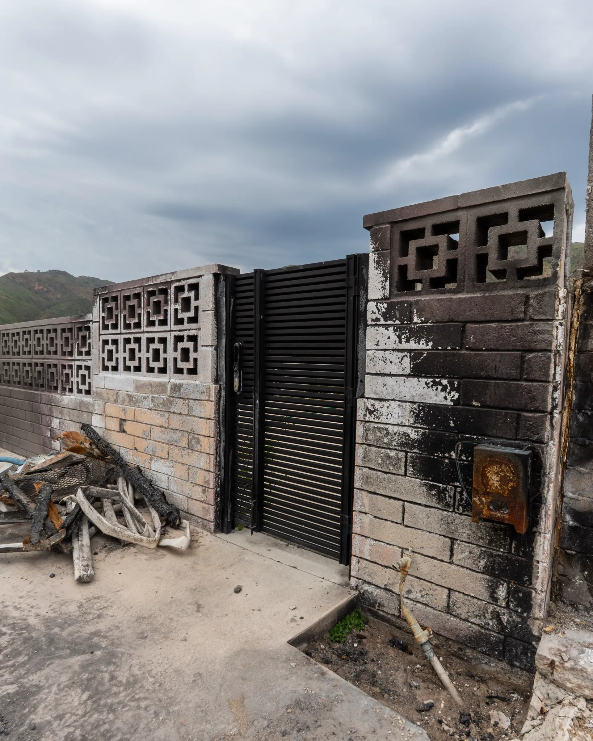 A black aluminum slat gate undamaged within a scorched concrete block wall, burned debris to one side
