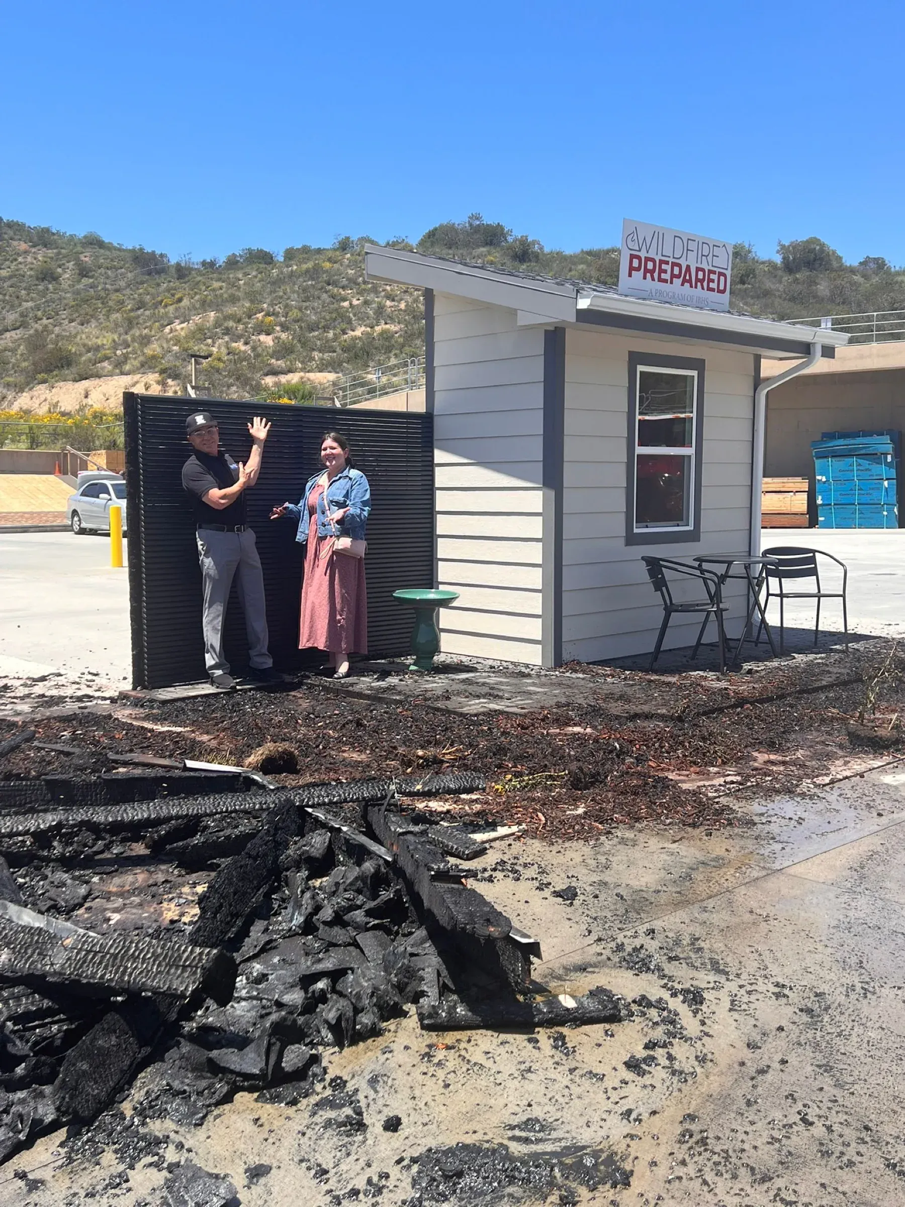 Two people standing at the undamaged Mulholland slat screen after the burn test, charred debris covering the ground in front of them