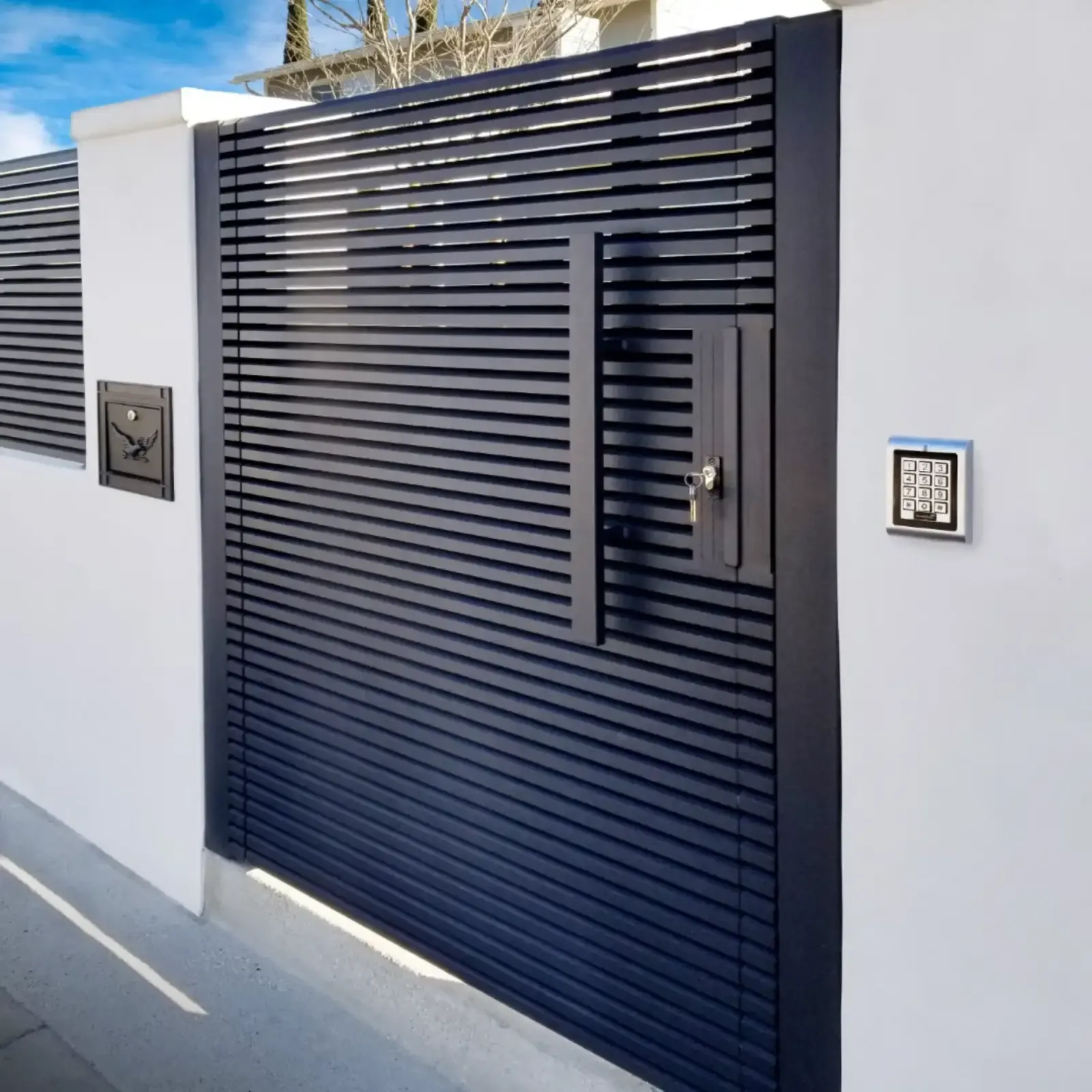 A dark slatted aluminum pedestrian gate set into a white boundary wall, with a keypad beside it.