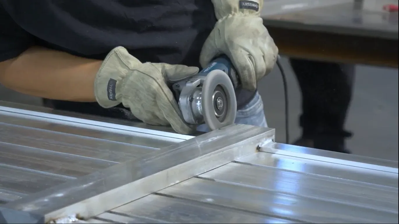 A gloved fabricator cutting an aluminum profile with an angle grinder on the factory floor
