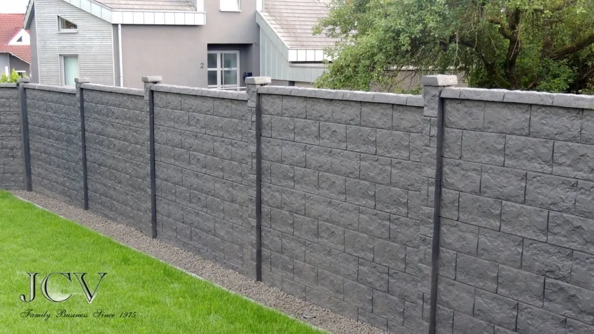 A dark grey concrete block wall with textured panels set between slim posts, along a garden boundary.