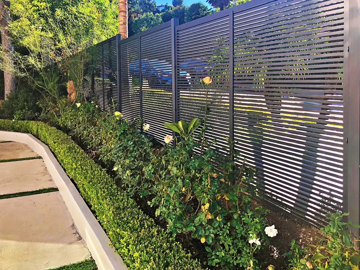 A dark horizontal-slat aluminum fence behind a bed of roses and a clipped hedge