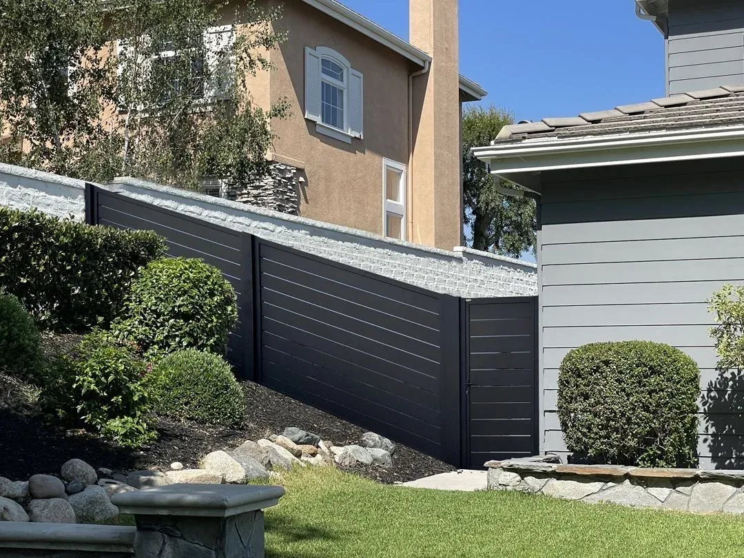 A dark aluminum fence on a raised bank between two houses, stepping up with the ground