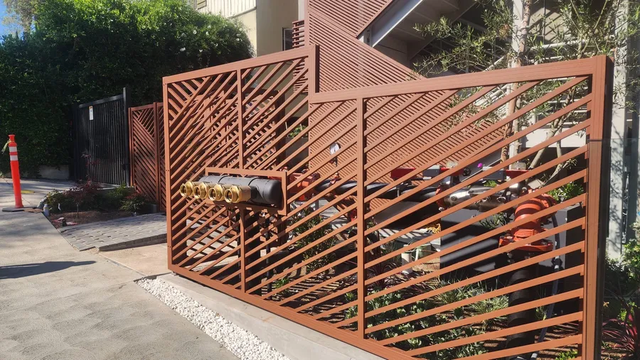 A bronze aluminum lattice screen enclosing utility risers and valves beside a driveway.
