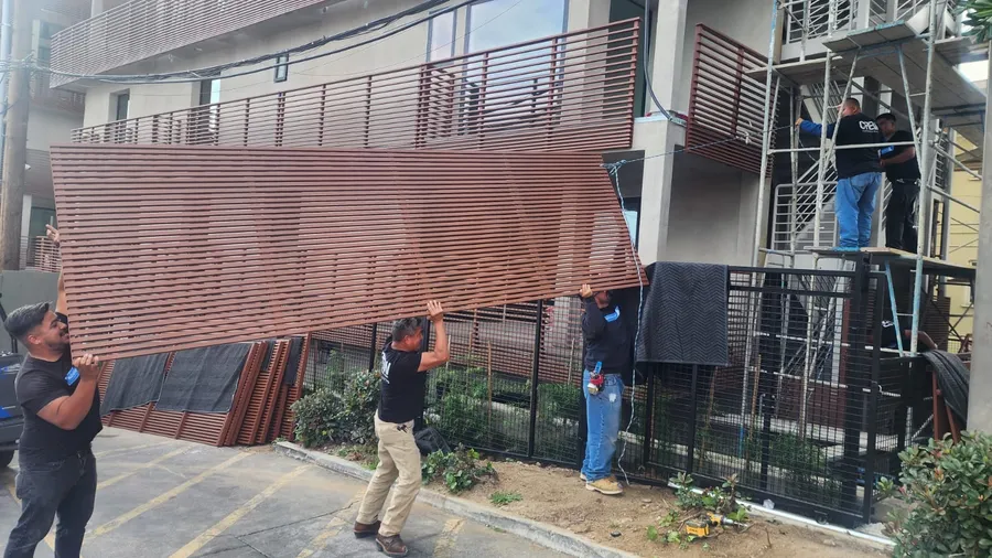 Five Mulholland installers carrying a full-height slat panel into position beside the building, with scaffolding behind them.