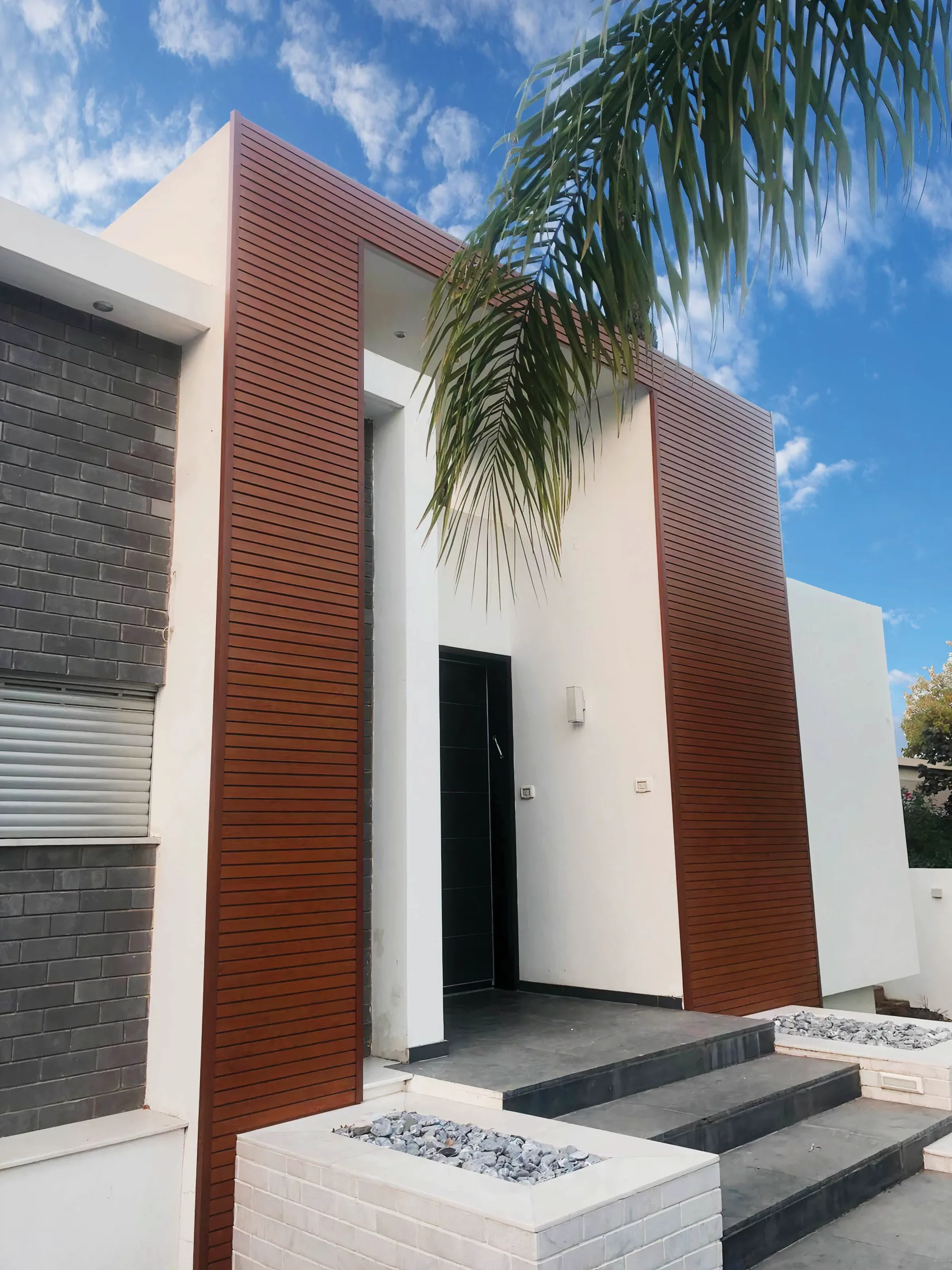 Two full-height wood-look aluminum panels flanking the white stucco entrance of a modern house, beside dark brickwork.