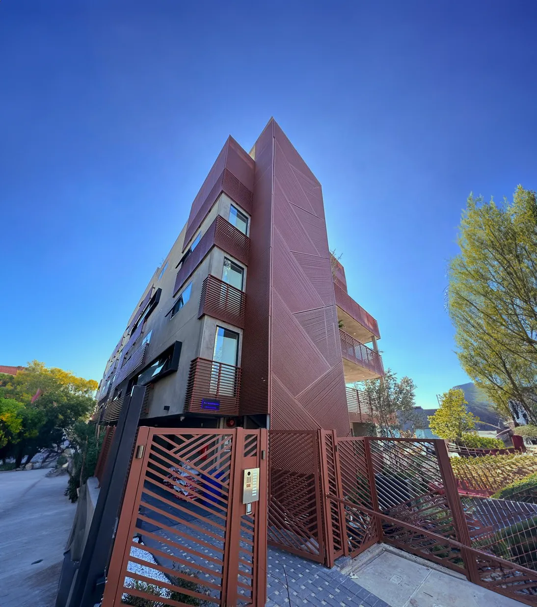 A four-storey apartment building on a corner, its stair core clad in patterned red-brown aluminum with matching balcony screens and street gates.